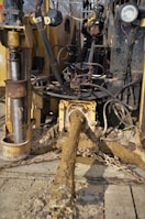 A muddy and rugged industrial machine with various cables and hydraulic components. It appears to be involved in heavy-duty work, potentially in construction or drilling. The setting seems outdoors with some greenery in the background.