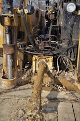A muddy and rugged industrial machine with various cables and hydraulic components. It appears to be involved in heavy-duty work, potentially in construction or drilling. The setting seems outdoors with some greenery in the background.