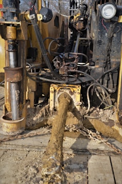 A muddy and rugged industrial machine with various cables and hydraulic components. It appears to be involved in heavy-duty work, potentially in construction or drilling. The setting seems outdoors with some greenery in the background.