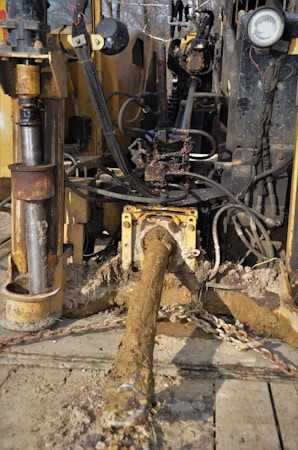 A muddy and rugged industrial machine with various cables and hydraulic components. It appears to be involved in heavy-duty work, potentially in construction or drilling. The setting seems outdoors with some greenery in the background.