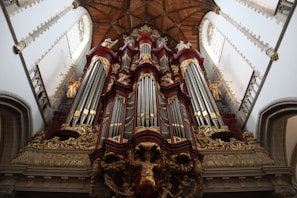 An ornate pipe organ is prominently displayed with intricate carvings and decorative elements. The structure features a series of large vertical pipes flanked by smaller decorative columns and statues. The setting appears to be a grand, historic church or cathedral interior with high ceilings and detailed architectural designs.