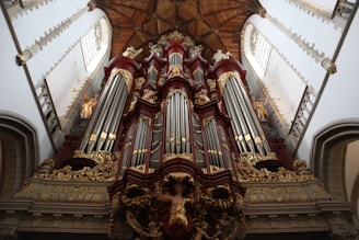 An ornate pipe organ is prominently displayed with intricate carvings and decorative elements. The structure features a series of large vertical pipes flanked by smaller decorative columns and statues. The setting appears to be a grand, historic church or cathedral interior with high ceilings and detailed architectural designs.