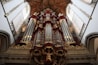 An ornate pipe organ is prominently displayed with intricate carvings and decorative elements. The structure features a series of large vertical pipes flanked by smaller decorative columns and statues. The setting appears to be a grand, historic church or cathedral interior with high ceilings and detailed architectural designs.