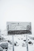 A heavily snow-covered sign with text in German stands amidst a snowy landscape. The sign is frosted over, obscuring some details, while snow blankets the surrounding area, including the ground and nearby foliage.