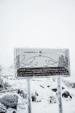 A heavily snow-covered sign with text in German stands amidst a snowy landscape. The sign is frosted over, obscuring some details, while snow blankets the surrounding area, including the ground and nearby foliage.