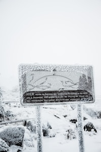 A heavily snow-covered sign with text in German stands amidst a snowy landscape. The sign is frosted over, obscuring some details, while snow blankets the surrounding area, including the ground and nearby foliage.