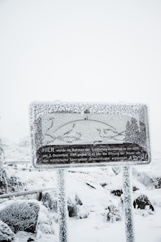 A heavily snow-covered sign with text in German stands amidst a snowy landscape. The sign is frosted over, obscuring some details, while snow blankets the surrounding area, including the ground and nearby foliage.