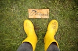 Two yellow rubber boots standing on grass with a small wooden sign between them that reads 'to Heaven' with a footprint illustration.