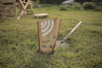 A wooden sign planted in a grassy area with the words 'NO WIFI ZONE' painted on it in a rustic style. In the background, there are some wooden structures and a blurred, natural setting that includes grass and bushes, providing an outdoor, rural vibe.