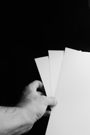 A hand holding three white sheets of paper against a dark background. The paper sheets are fanned out, and the scene is captured in black and white.