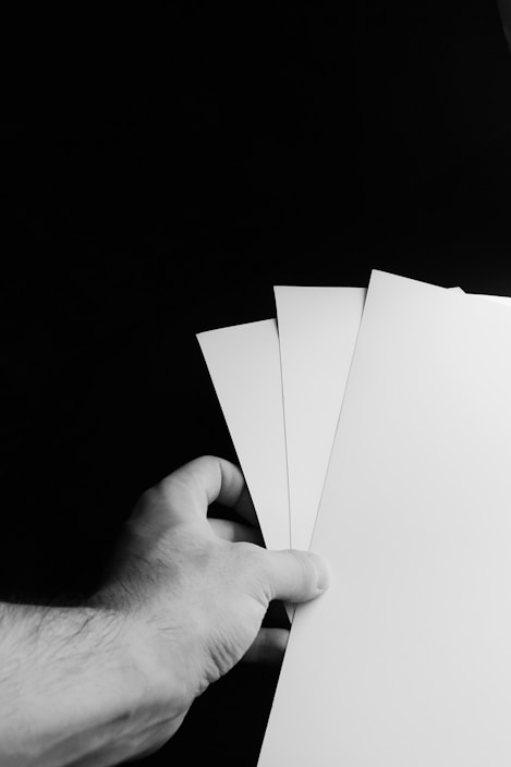 A hand holding three white sheets of paper against a dark background. The paper sheets are fanned out, and the scene is captured in black and white.