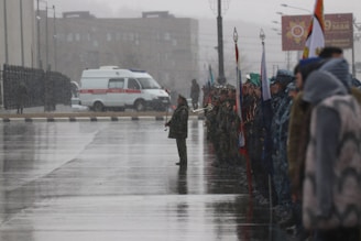 A line of uniformed individuals, possibly military personnel, stand in formation holding flags. An officer stands facing them in the rain. An ambulance is parked nearby, and the scene is set against a backdrop of urban buildings. The overcast sky and rain create a somber atmosphere.
