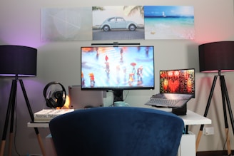 A modern workspace setup featuring a large monitor, a laptop, and a pair of lamps on either side of the desk. The monitor displays an image of people walking with umbrellas on a rainy street. The walls are decorated with three framed pictures, including a car and a beach scene. A pair of headphones rests on a stack of books, with 'Atomic Habits' visible. The setup is organized and illuminated with soft lighting, creating a cozy environment.