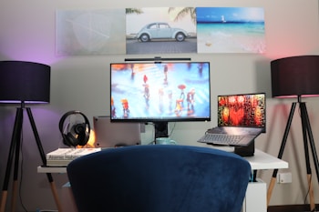 A modern workspace setup featuring a large monitor, a laptop, and a pair of lamps on either side of the desk. The monitor displays an image of people walking with umbrellas on a rainy street. The walls are decorated with three framed pictures, including a car and a beach scene. A pair of headphones rests on a stack of books, with 'Atomic Habits' visible. The setup is organized and illuminated with soft lighting, creating a cozy environment.