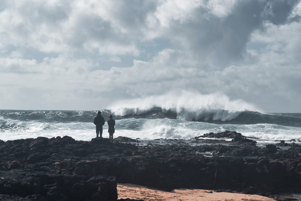 2 person standing on rock near sea waves under cloudy sky during daytime