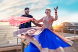 A couple dressed in traditional Indian attire, both wearing sunglasses, are dancing joyfully on a rooftop with a scenic ocean view. The woman is wearing a vibrant blue and pink lehenga, while the man is in a blue sherwani. The background shows a coastal cityscape under a clear blue sky, with the sun setting near a large domed building.