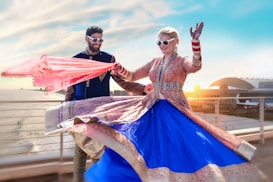 A couple dressed in traditional Indian attire, both wearing sunglasses, are dancing joyfully on a rooftop with a scenic ocean view. The woman is wearing a vibrant blue and pink lehenga, while the man is in a blue sherwani. The background shows a coastal cityscape under a clear blue sky, with the sun setting near a large domed building.