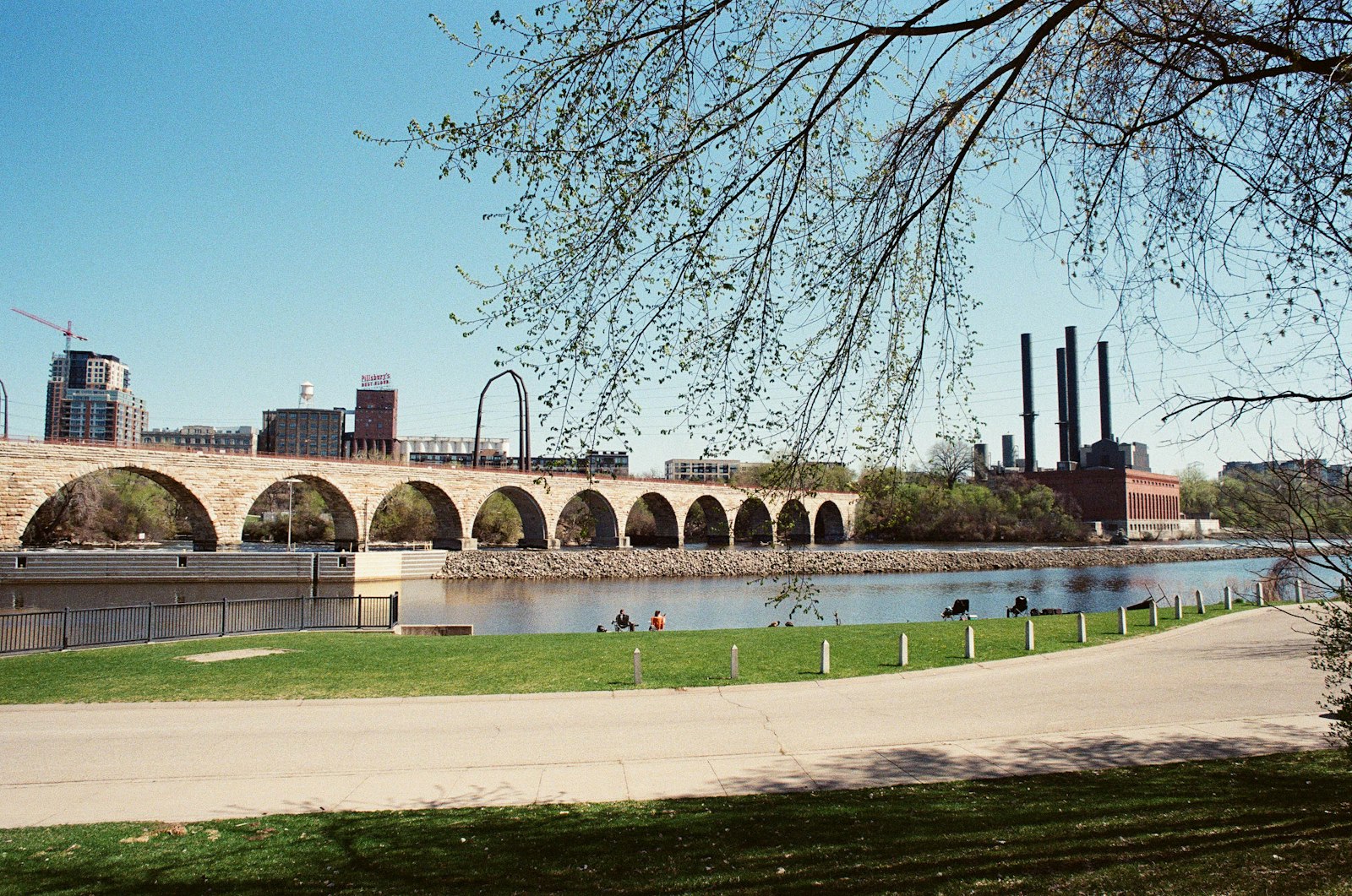 A paved riverside path curving past a lawn with the arches of the Stone Arch Bridge and old mill buildings behind