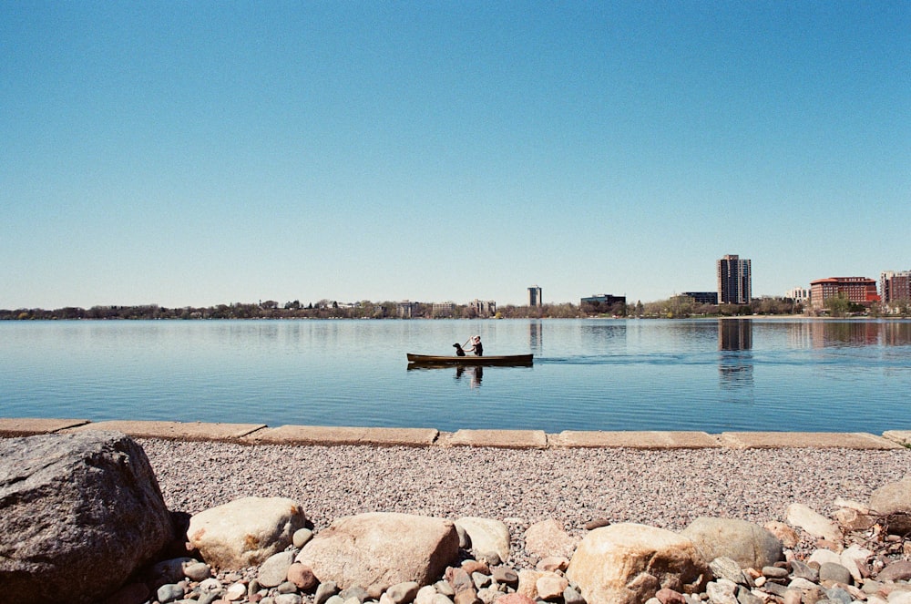 Persona con camisa negra de pie sobre una roca marrón cerca del cuerpo de agua durante el día