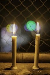 Two lit candles with wax drips are placed on a surface in front of a wire mesh, with soft, out-of-focus colorful lights in the background giving a warm ambiance.