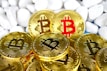 Several gold-colored coins featuring the Bitcoin logo are arranged in a pile against a backdrop of smooth white stones. One coin stands out with a red Bitcoin symbol.