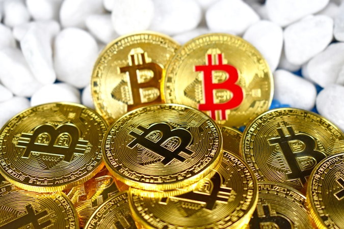 Several gold-colored coins featuring the Bitcoin logo are arranged in a pile against a backdrop of smooth white stones. One coin stands out with a red Bitcoin symbol.