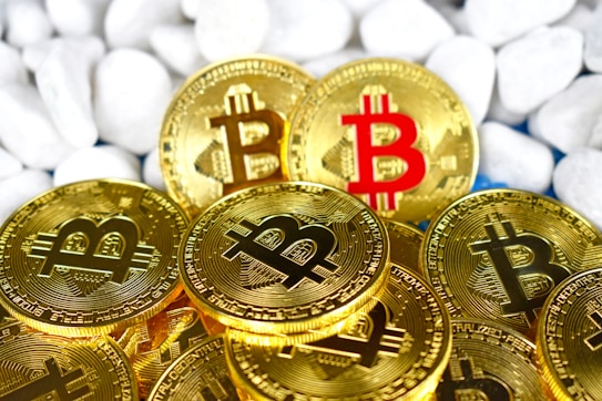 Several gold-colored coins featuring the Bitcoin logo are arranged in a pile against a backdrop of smooth white stones. One coin stands out with a red Bitcoin symbol.