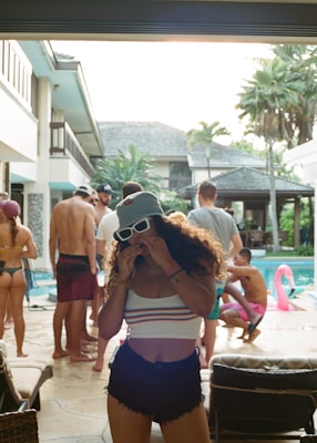 A group of people are gathered around a swimming pool at what appears to be a pool party. Some individuals are standing while others are sitting or crouching near the water. The setting is in a residential area with palm trees and a house in the background. One person in the foreground is wearing sunglasses, a hat, and casual summer attire, eating something.