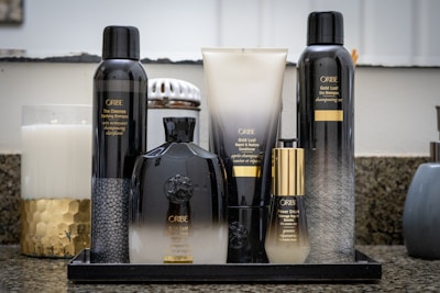 A neatly arranged set of Oribe hair care products sits on a granite countertop. The collection includes bottles and tubes of various sizes, predominantly black and gold in color. A large white candle and a decorative jar can be seen in the background, adding a touch of elegance to the setting.