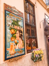 A vintage-style poster on a wall depicts a woman with elaborate hair accessories, surrounded by oranges and foliage. The text on the poster promotes 'Agua de Valencia', with 'Nuestra Especialidad' written below. The scene is complemented by a nearby window with bars and a vibrant floral arrangement, adding a traditional and cheerful ambiance.