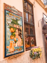 A vintage-style poster on a wall depicts a woman with elaborate hair accessories, surrounded by oranges and foliage. The text on the poster promotes 'Agua de Valencia', with 'Nuestra Especialidad' written below. The scene is complemented by a nearby window with bars and a vibrant floral arrangement, adding a traditional and cheerful ambiance.