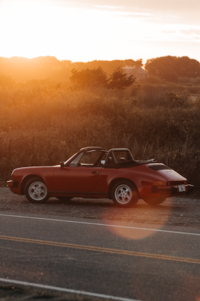 A classic red convertible car is parked on the side of a road during sunset. The scene is set in a rural area with a backdrop of trees and fields bathed in warm, golden light. The road is empty, and the sun's rays create a lens flare, adding a nostalgic feel to the image.