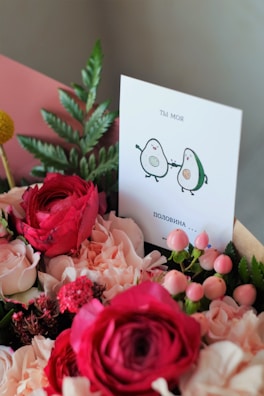 A vibrant bouquet of flowers, including red and light pink roses, is accompanied by a card. The card features an illustration of two cartoon avocados holding hands, with text written in a Cyrillic script.