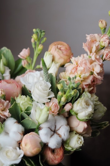 A bouquet of flowers featuring a mix of roses, cotton flowers, and various buds in soft pastel colors, arranged with green leaves. The delicate flowers are predominantly white and light pink, giving a fresh and elegant appearance.