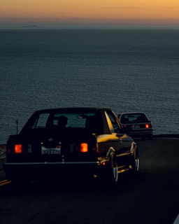 Two cars driving along a coastal road during sunset, with the ocean in the background and a horizon tinged with orange and yellow hues.