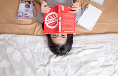 A person lying on a bed is reading a book titled 'Unmarketing' with a bright red cover. The bed has a beige and white pattern blanket, and there are a couple of other books nearby, one with an illustrated cover and another with a plain white cover.