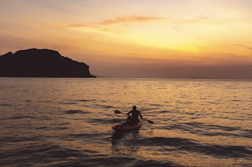 A person is kayaking on calm waters during a sunset or sunrise. The silhouette of the kayaker is prominent against the golden hues of the sky and the reflection on the water. In the background, there is a large, dark silhouette of a mountain or an island.