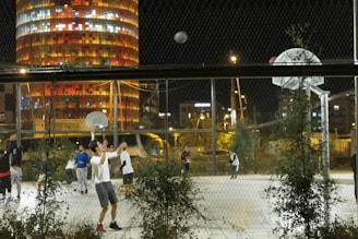 A nighttime basketball game is taking place on an outdoor court surrounded by a chain-link fence. Several young men are actively engaged in the game, and a few are seen shooting and moving around the court. In the background, a tall, brightly lit cylindrical building with colorful windows illuminates the night sky.