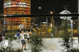 A nighttime basketball game is taking place on an outdoor court surrounded by a chain-link fence. Several young men are actively engaged in the game, and a few are seen shooting and moving around the court. In the background, a tall, brightly lit cylindrical building with colorful windows illuminates the night sky.
