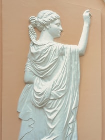 A relief sculpture of a classical figure, depicted in profile, wearing flowing robes and with intricately braided hair. The figure raises one arm, while the background of the sculpture is a muted peach color.