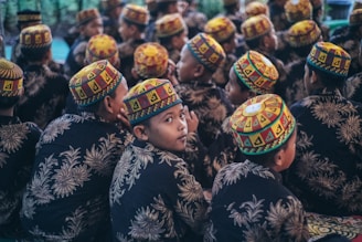 A group of children is seated facing away, wearing colorful, patterned skullcaps and dark clothing with floral designs. One child looks back at the camera with a curious expression.