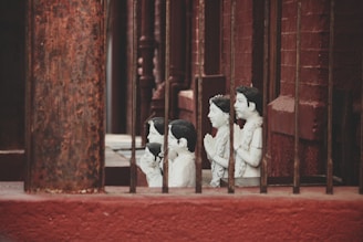 A series of small, white statues depicting people with hands in a prayer position are positioned behind metal bars. The statues appear to be placed along a red brick wall, creating a contrast with their pale color.