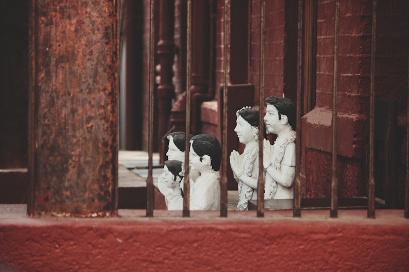 A series of small, white statues depicting people with hands in a prayer position are positioned behind metal bars. The statues appear to be placed along a red brick wall, creating a contrast with their pale color.