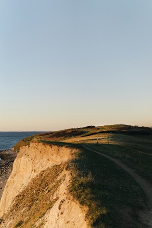 A coastal landscape featuring a path winding along grassy cliffs. The cliffs overlook a rocky shoreline which meets a calm sea. The sky is clear with a soft gradient from light blue to subtle golden hues, suggesting early morning or late afternoon sunlight.