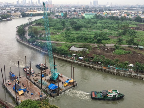 A river scene featuring a construction site on a floating platform with cranes and equipment. There is a barge or tugboat maneuvering nearby, and a lush green landscape with trees along the riverbank. Background includes urban infrastructure, buildings, and roads, with a city skyline in the distance.