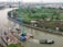 A river scene featuring a construction site on a floating platform with cranes and equipment. There is a barge or tugboat maneuvering nearby, and a lush green landscape with trees along the riverbank. Background includes urban infrastructure, buildings, and roads, with a city skyline in the distance.