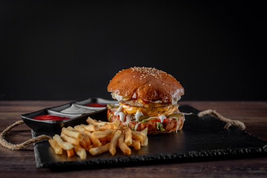 A juicy cheeseburger with sesame seed bun is presented on a black slate board, accompanied by a side of crispy French fries. The burger is topped with melted cheese, lettuce, and sauces. Two containers hold ketchup and mayonnaise, adding to the appetizing presentation.
