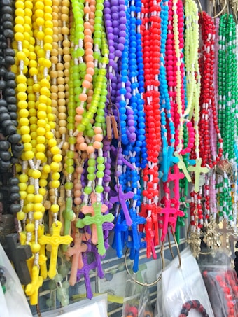 A vibrant display of colorful rosary beads hangs in rows. Each string features beads in shades of yellow, green, purple, blue, red, and pink, with matching crucifixes at the bottom. The arrangement creates a visually striking and diverse assortment.