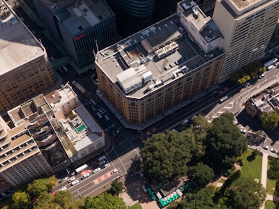 An aerial view of a city block with tall, rectangular buildings. The streets form a grid pattern with several vehicles visible on the road. One building has a rooftop pool, and there are large patches of greenery with trees and a park area below.