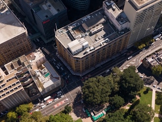 An aerial view of a city block with tall, rectangular buildings. The streets form a grid pattern with several vehicles visible on the road. One building has a rooftop pool, and there are large patches of greenery with trees and a park area below.